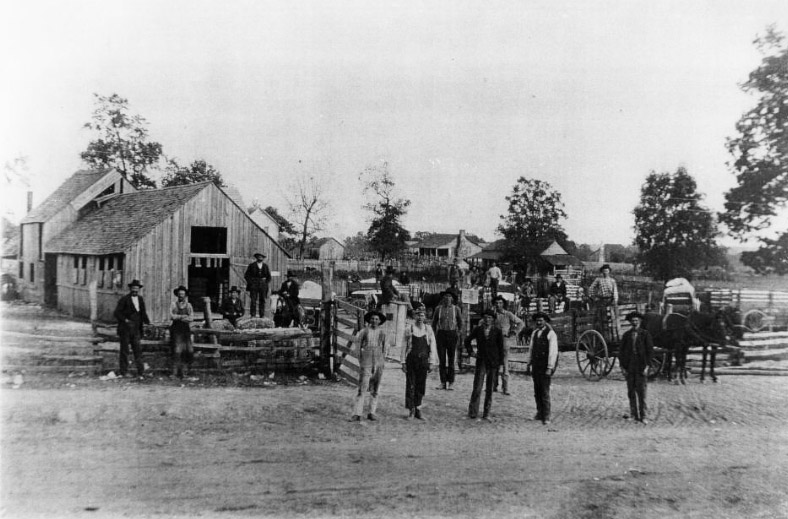 White men with horse drawn wagon standing outside cotton gin buildings with houses in the background
