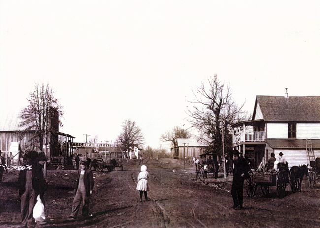 White men in overalls and little girl in white dress standing in dirt road with stores and trees on both sides