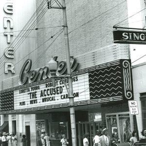 passersby walking on sidewalk at multistory theater building with "The Accused" on its marquee