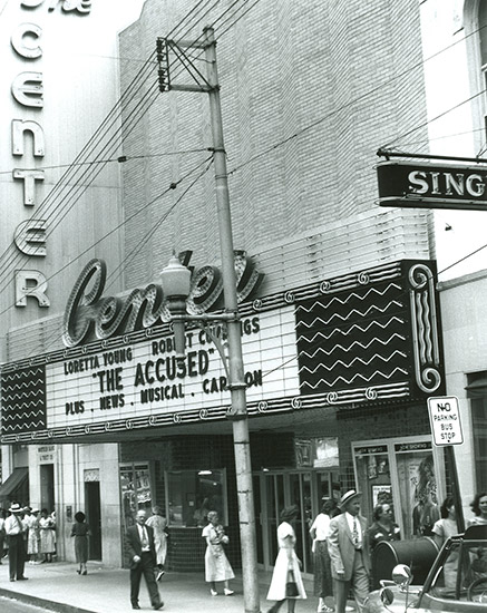 passersby walking on sidewalk at multistory theater building with "The Accused" on its marquee