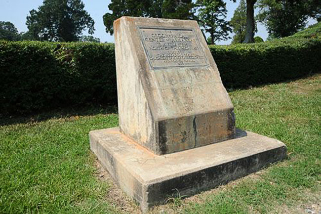 Concrete monument with base and plaque and hedge behind it