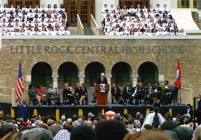 white man in suit and red tie on dais with other individuals, white and African American, in front of brick building labeled "Little Rock Central High School" before a large crowd