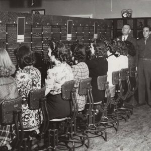 Group of white women sitting at switchboard while white men and woman watch them work