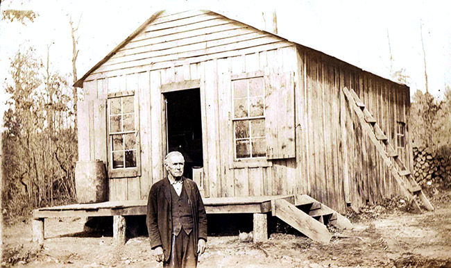 Older white man in suit standing outside single-story building