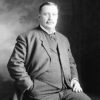 White man with mustache standing in suit and tie