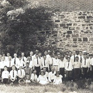 Group of white men women and children posing before stone wall with tree