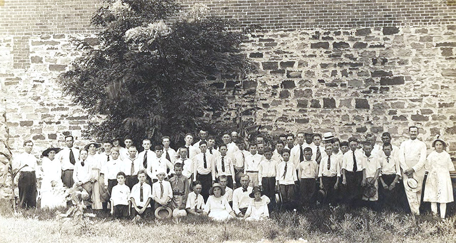 Group of white men women and children posing before stone wall with tree