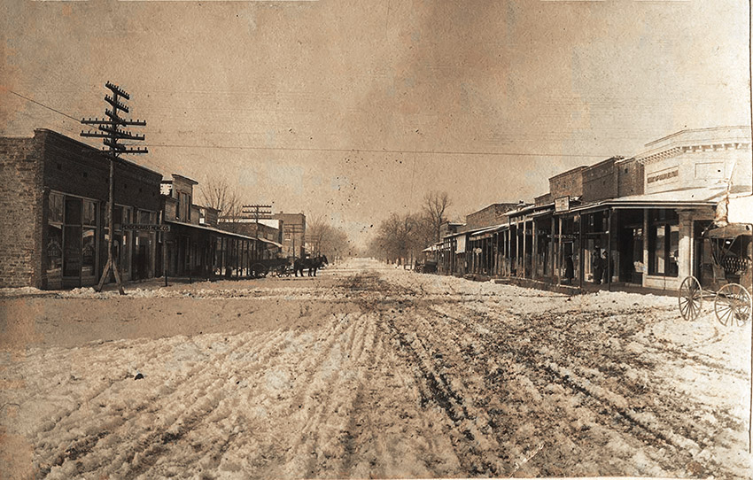 Dirt town street with storefronts on both sides and horses in the middle distance