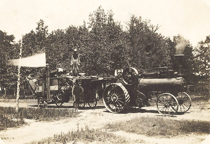 Three men posing on dirt road with large steam-powered tractor attached to heavy equipment with trees in background