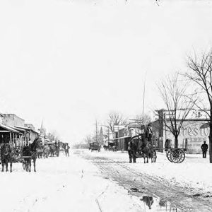 Horse drawn wagons on snow covered street between rows of one and two story brick buildings