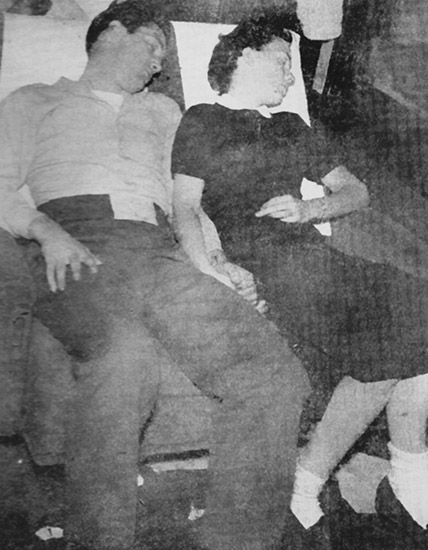 White man in shirt and pants and woman in a dark dress holding hands and sleeping in train car