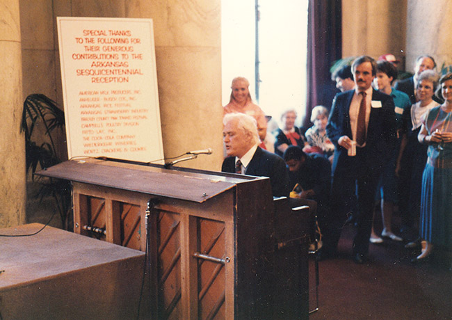 Old white man in suit playing piano and singing for a mixed crowd