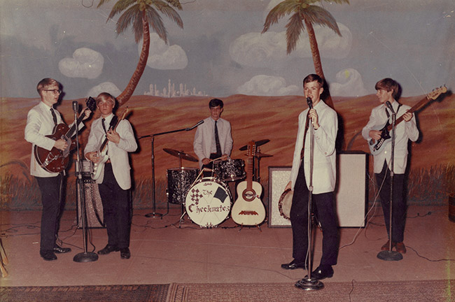 Five young white men in white jackets and black pants playing instruments on stage in front of a backdrop with palm trees