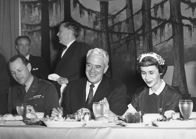 White men in suits and military uniform and woman eating a banquet with fake forest scene behind them