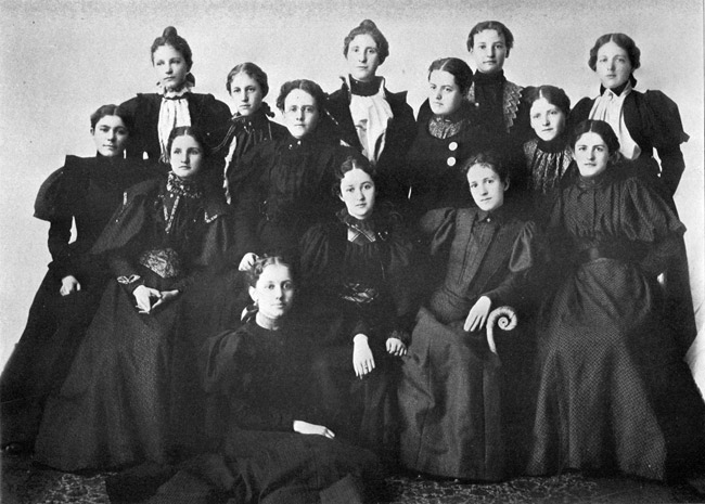 Group of more than a dozen white women posing together in black dresses and pulled-back hair