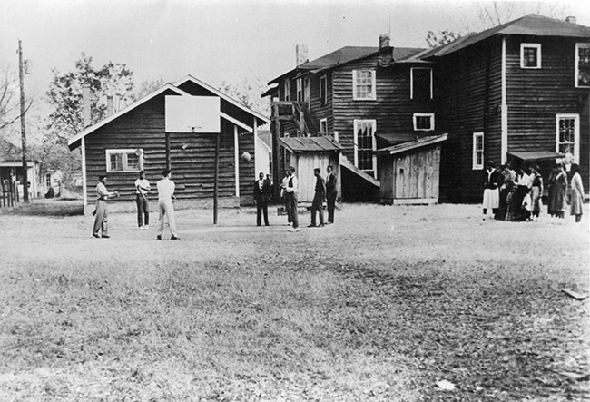 young African-American men playing basketball outside with school buildings in the background and young African American women watching