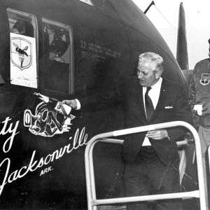 Older white man in suit and tie holding a champagne bottle against "City of Jacksonville" plane while a smiling man in military uniform watches