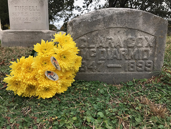 Yellow flowers with "I Voted" stickers next to small gravestone in cemetery