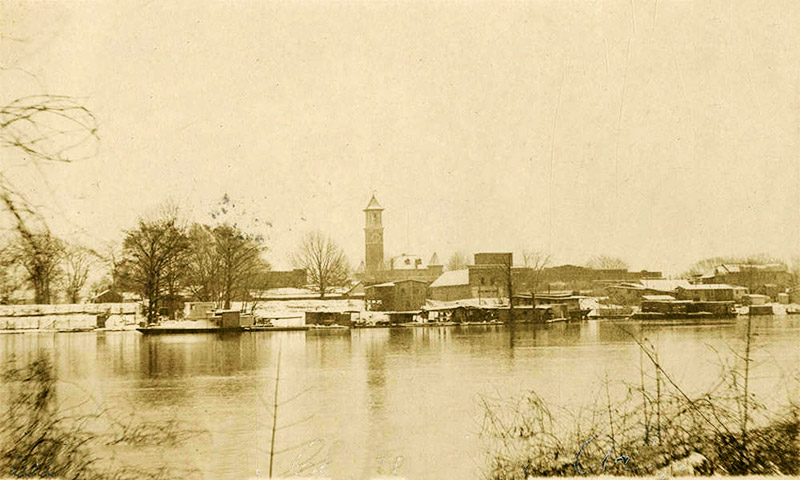View of town buildings and multistory building with clock tower as seen from across river