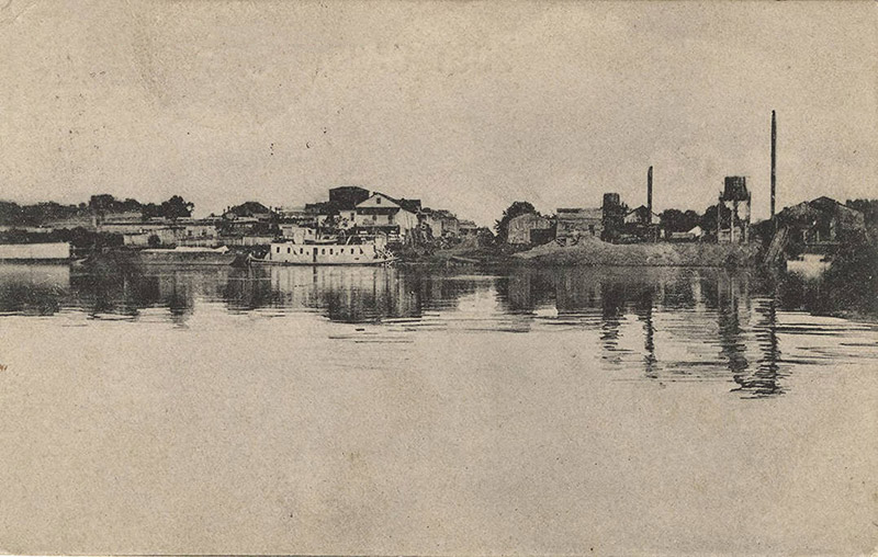 View from across river of town buildings and steamboat floating alongside the shore