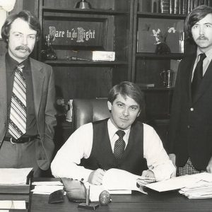 Two white men standing and white man sitting at desk in office