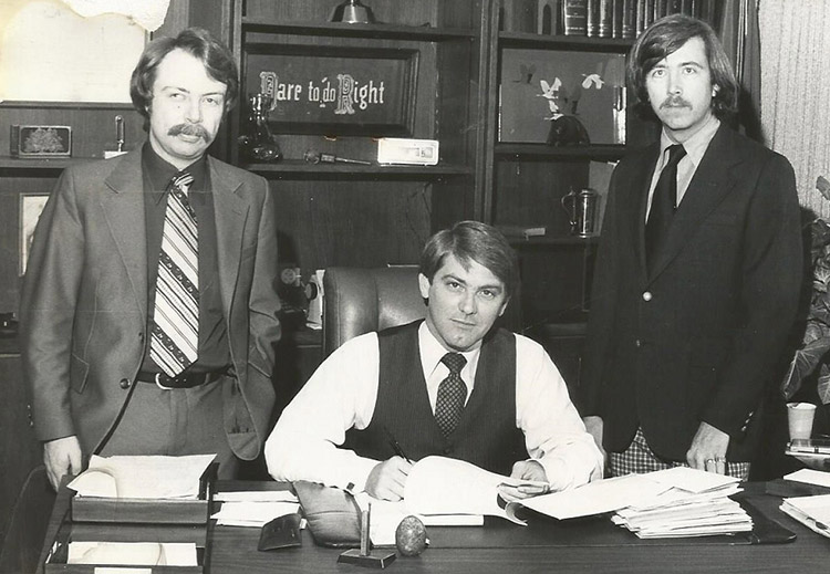Two white men standing and white man sitting at desk in office