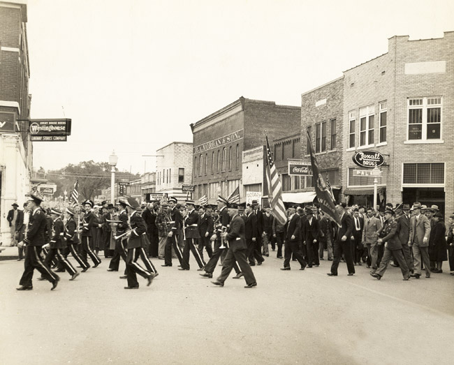 Downtown parade with marching band soldiers spectators brick buildings "rexall drugs westinghouse coca cola."
