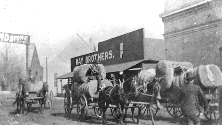 Men transporting cotton bales with horse-drawn wagons on town road