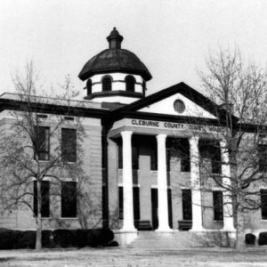 Multistory classical brick building with dark trim and "Cleburne County Court House" sign on the front with steeple dome trees hedges and large lawn