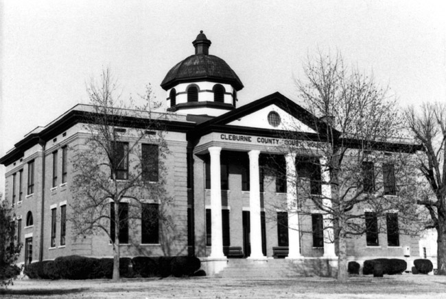 Multistory classical brick building with dark trim and "Cleburne County Court House" sign on the front with steeple dome trees hedges and large lawn
