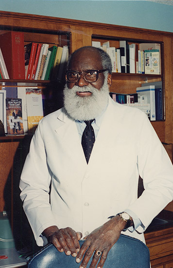 African-American man with bushy white beard in suit and tie standing in front of bookcase in office