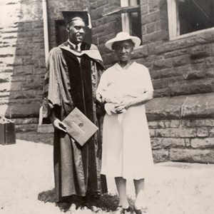 Young African-American man in graduation robes and cap with older African-American woman outside brick building