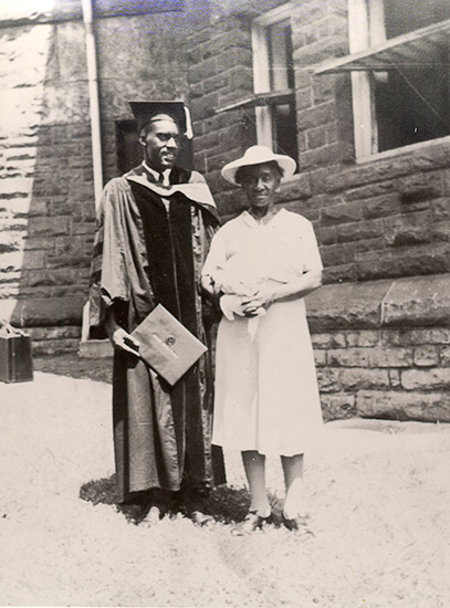 Young African-American man in graduation robes and cap with older African-American woman outside brick building