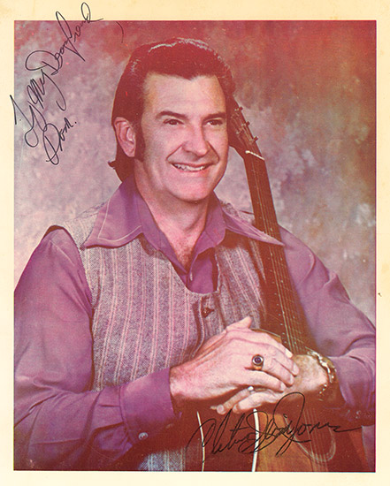 signed photo with white man in purple shirt and vest holding an acoustic guitar