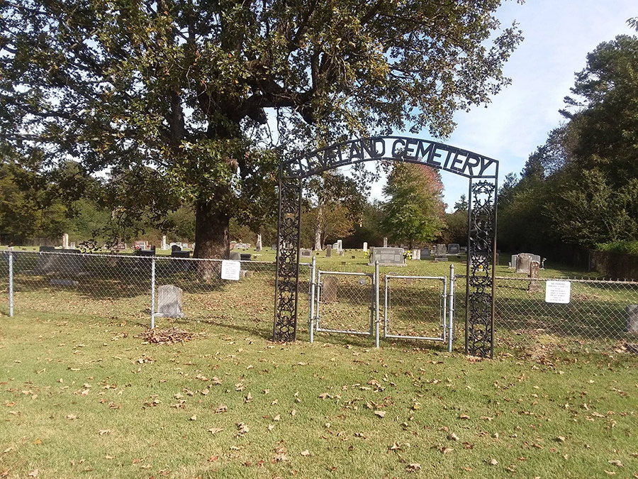 Iron "Cleveland Cemetery" arch with gravestones and tree beyond fence and gate