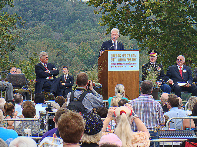 White man with white hair speaking into a microphone at lectern to a crowd with white men in suits and military officer on stage