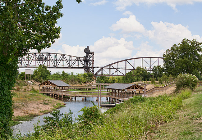 Steel truss bridge over river behind wooden pavilions connected by wooden walking bridge