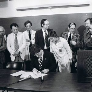 White man in suit sitting at classroom table with row of white men in suits and white woman in lab coat