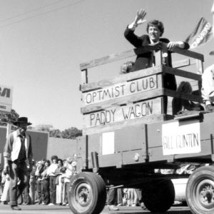 Man in suit waving at the crowd riding in a parade float labeled the "optimist club paddy wagon"