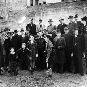 Group of white men and women in hats and suits with white children watching white boy use a shovel
