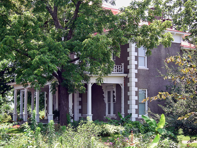 Two-story house with porch and decorative brick corners and tower behind a tree with gardens