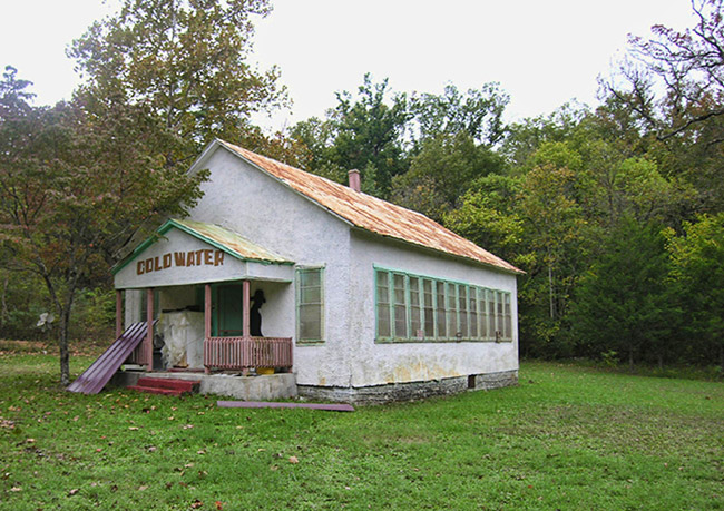 rundown white building with green trim with "Cold Water" painted above the entrance and a black silhouette on the porch