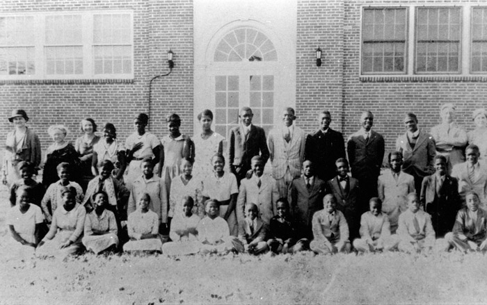 Group of children and young African-American men and women with white teachers in front of brick building with arched windows