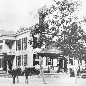Horse tied to a post outside two-story building with covered porch and balcony
