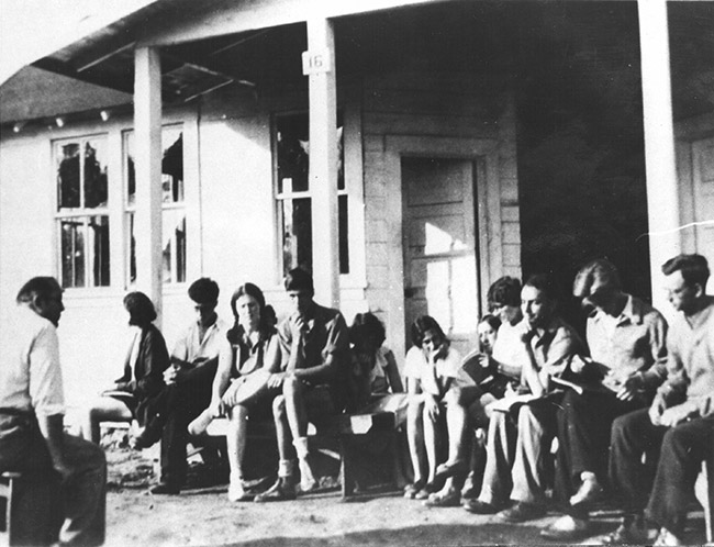 White man sitting in front of white students on porch of wooden building