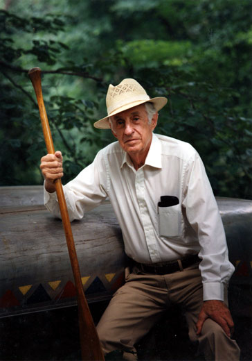 Old white man in hat holding a paddle posing with canoe and trees in the background
