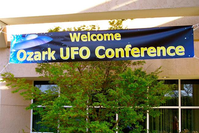 "Welcome Ozark U.F.O. Conference" banner on building with square panel windows and tree