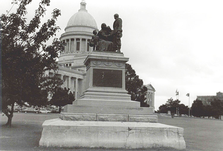 Front view of stone monument with statues atop pedestal with Capitol building behind it