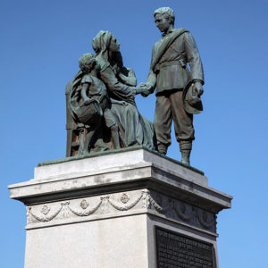 Statue of woman and child seated holding man's hand on pedestal with plaque on it