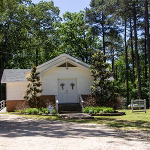 Church building with white siding and brick foundation on gravel parking lot with sign